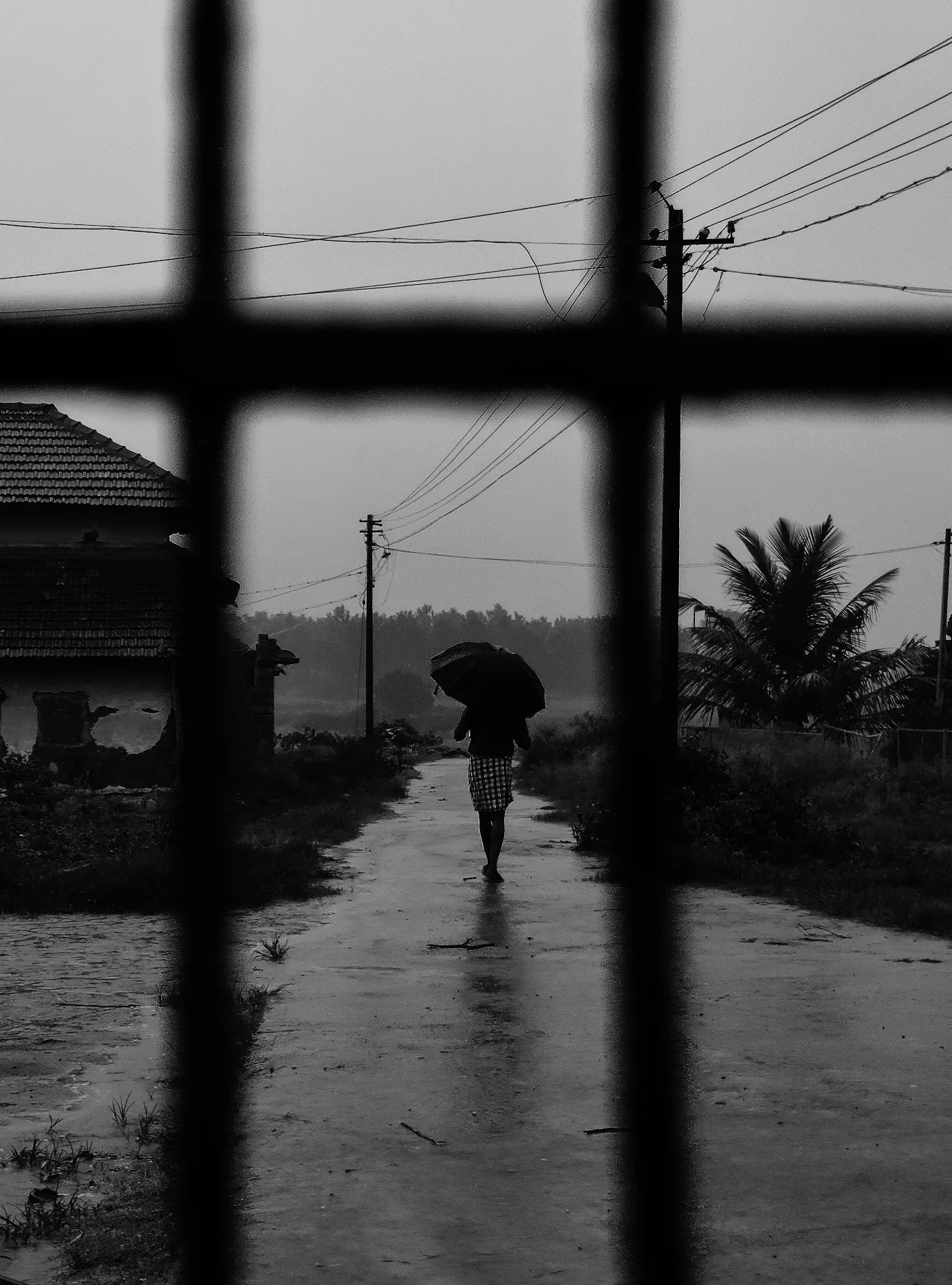 A rainy street seen through window bars, someone walking under an umbrella.